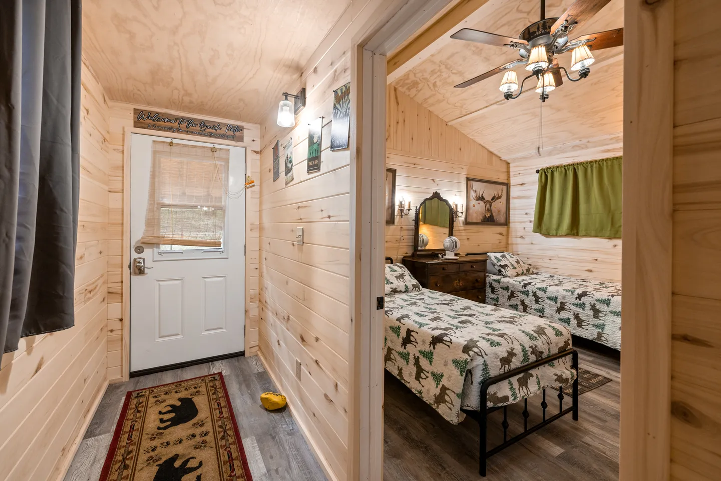 Entry hallway with light wood-paneled walls leading to an exterior door, and an open doorway showing a bedroom with two twin beds.