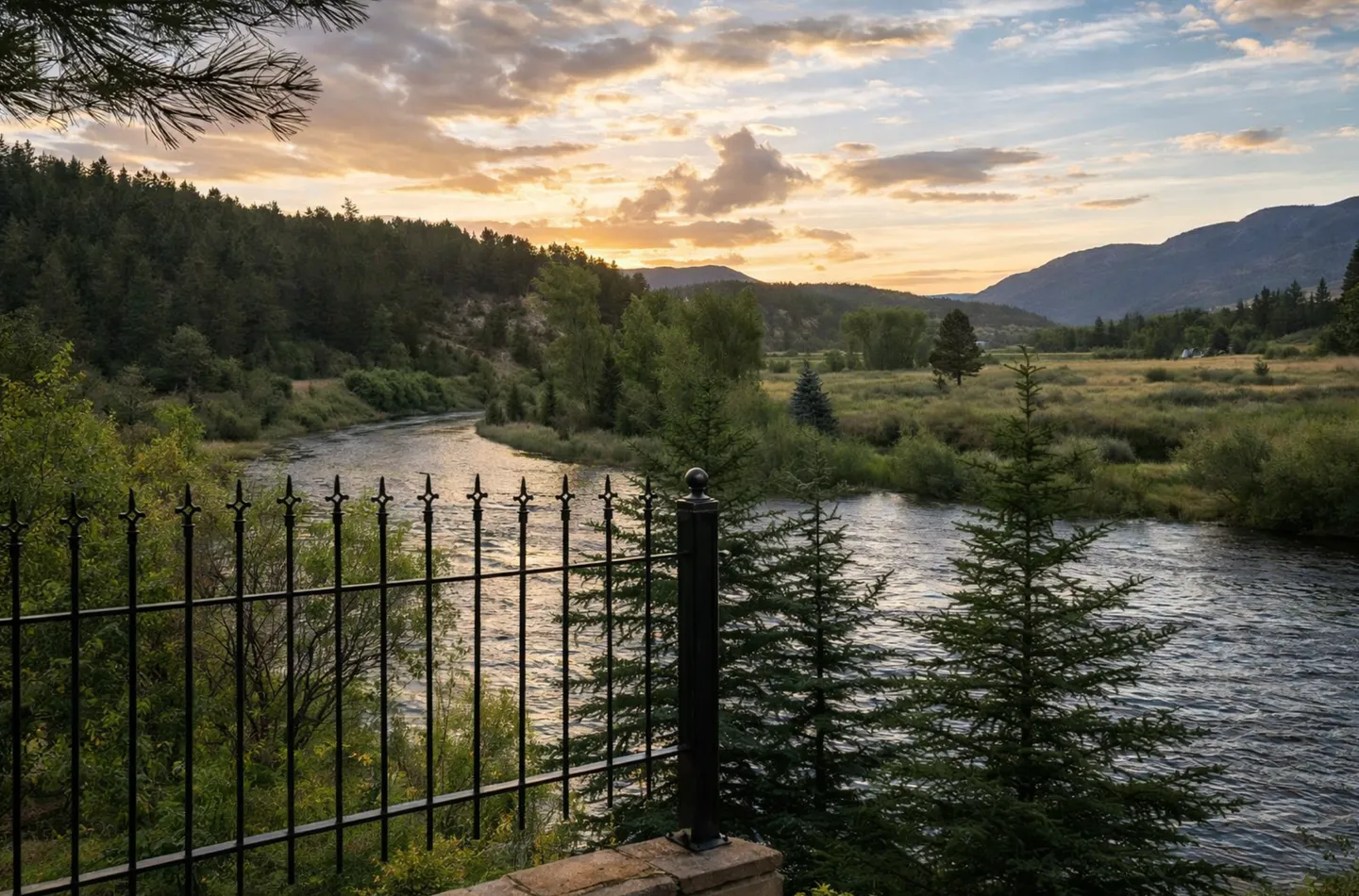 A view of a winding river surrounded by evergreen trees and mountains at sunset, framed by a black metal railing in the foreground.