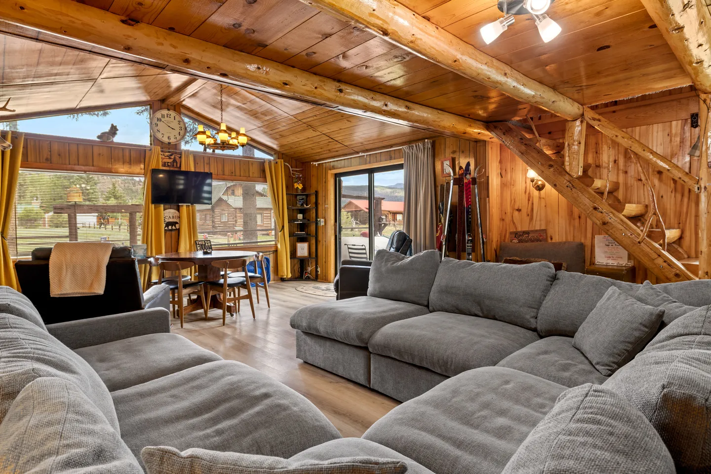 Living room with wood paneled walls, log beams, large grey sectional sofa, and windows looking out onto neighboring cabins.