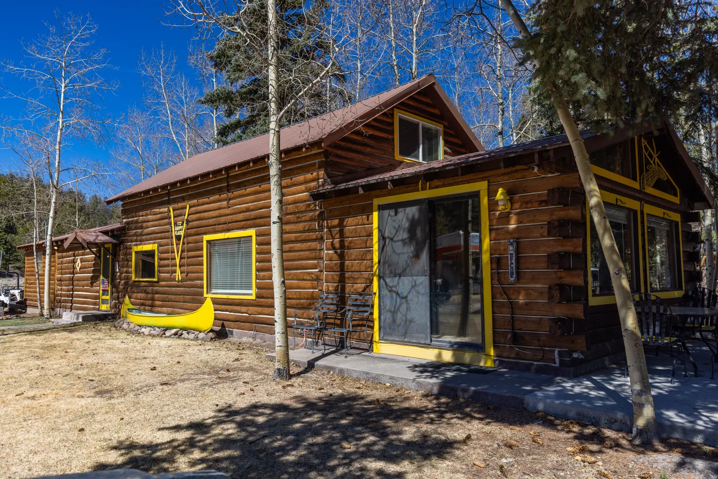 Exterior of a rustic log cabin with yellow window trim, a yellow decorative canoe, and patio seating under tall bare trees and a clear blue sky.