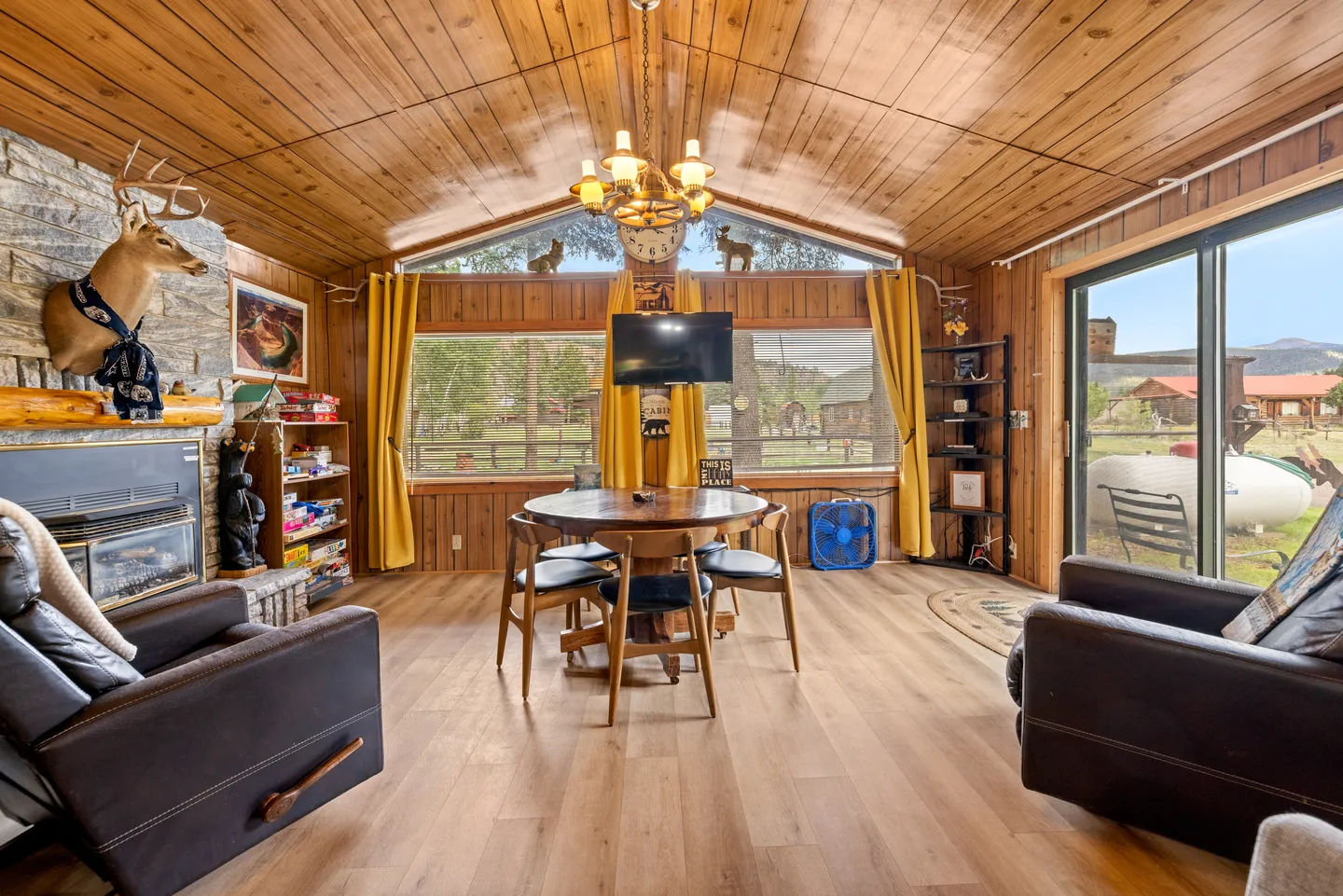 A rustic living room with wood-paneled walls and ceiling, a round dining table, leather armchairs, a fireplace, and large windows looking outside.