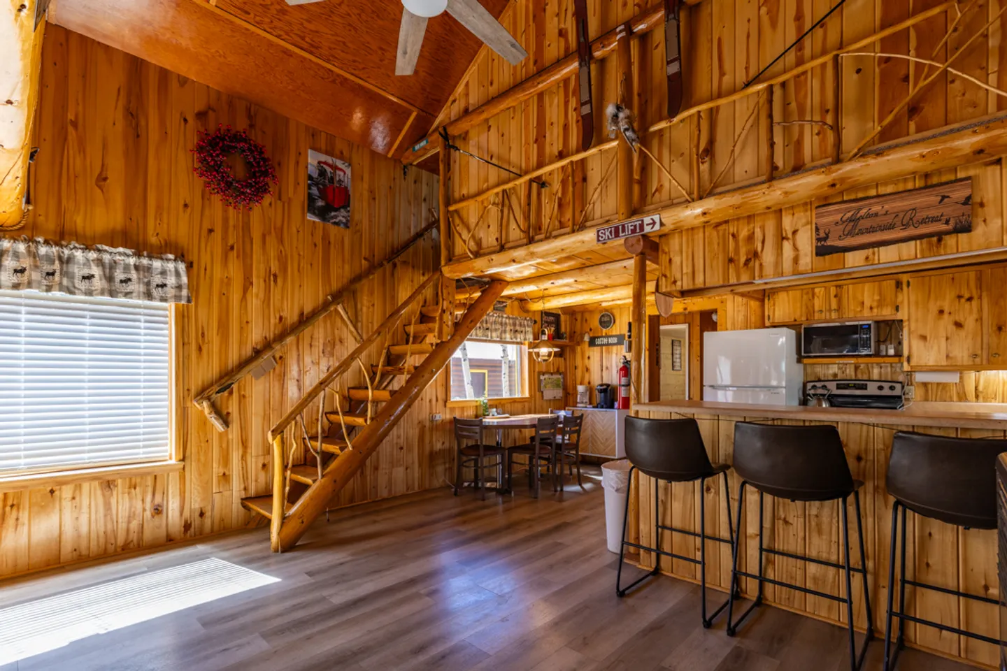 Open-concept cabin interior with knotty pine walls, wood staircase leading to a loft, kitchen bar with barstools, and dining table.