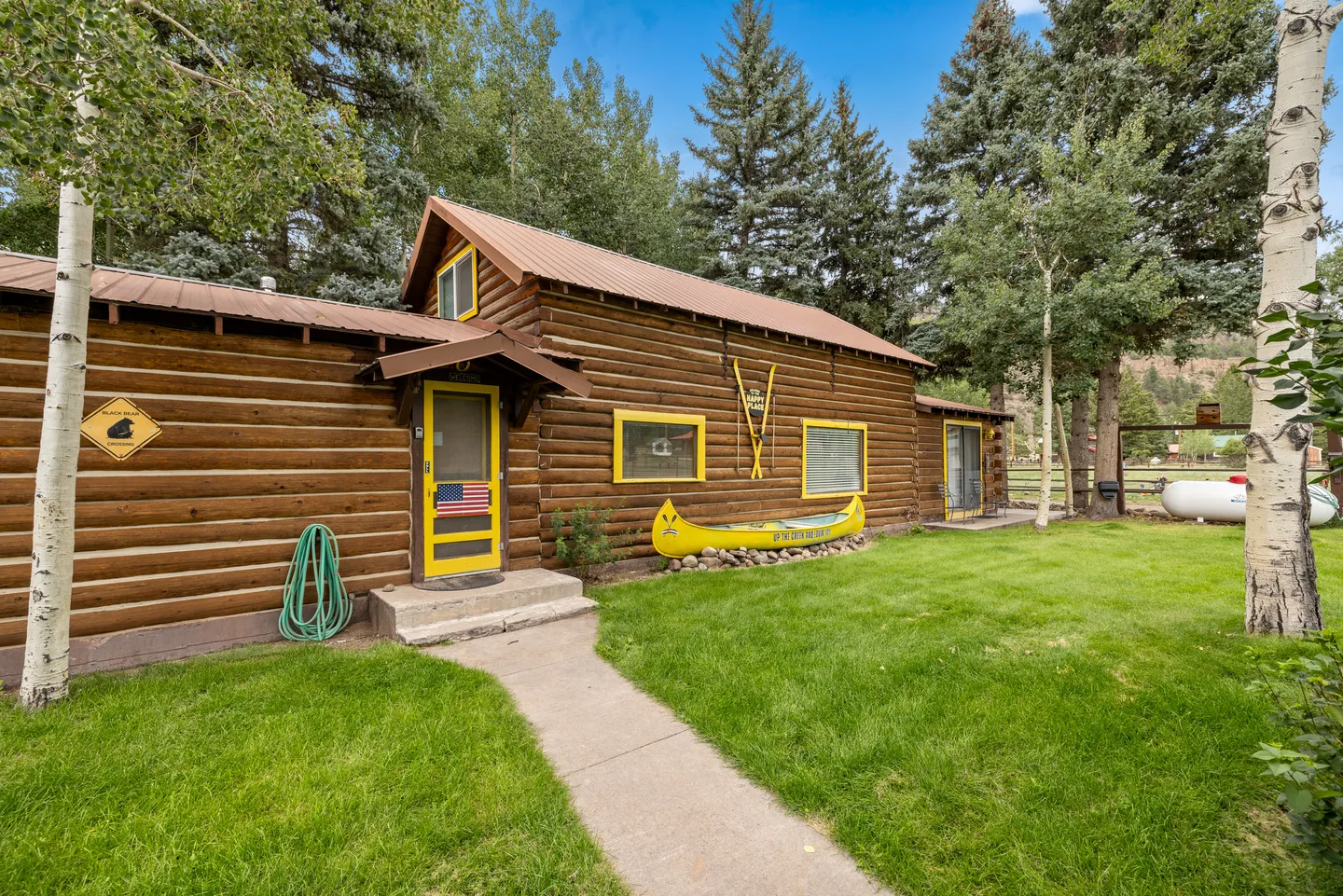 Exterior of a rustic log cabin with a yellow door, yellow-trimmed windows, a decorative yellow canoe, and a green lawn surrounded by tall trees.