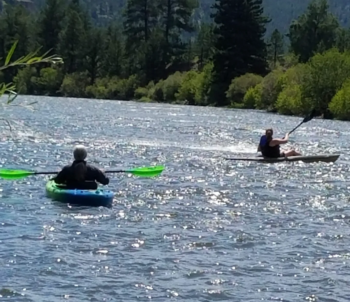 Two people kayaking on a sunlit river bordered by trees and hills.