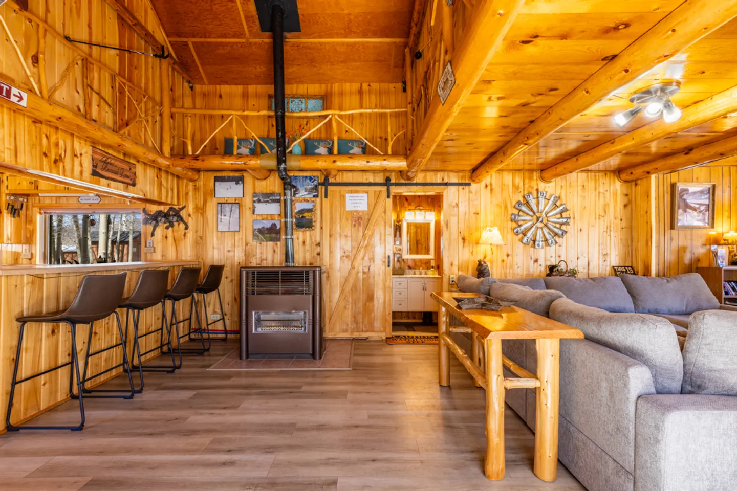 Rustic wooden living area with a bar and stools, a freestanding fireplace stove, a gray sectional sofa, and a doorway to a bathroom.