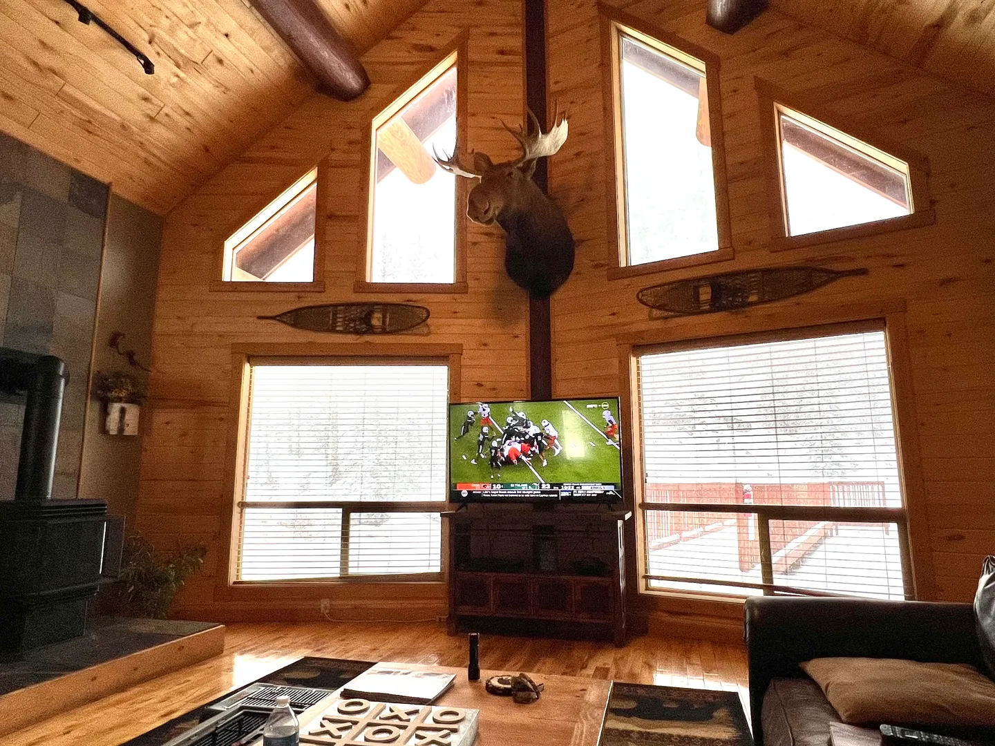 Living room with wood-paneled walls and ceiling, a television, large windows, a wood stove, and a mounted moose head.