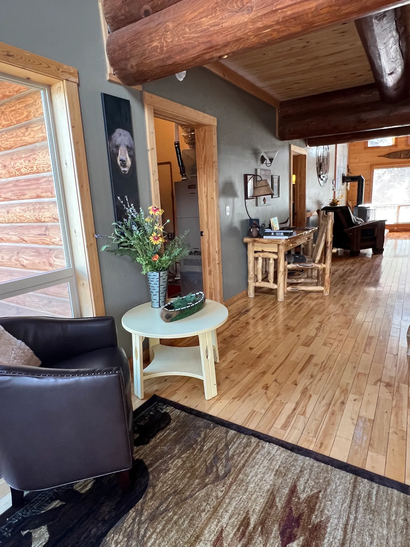 A rustic cabin hallway and sitting area featuring exposed log ceiling beams, wood flooring, a leather armchair, and a small wooden desk.