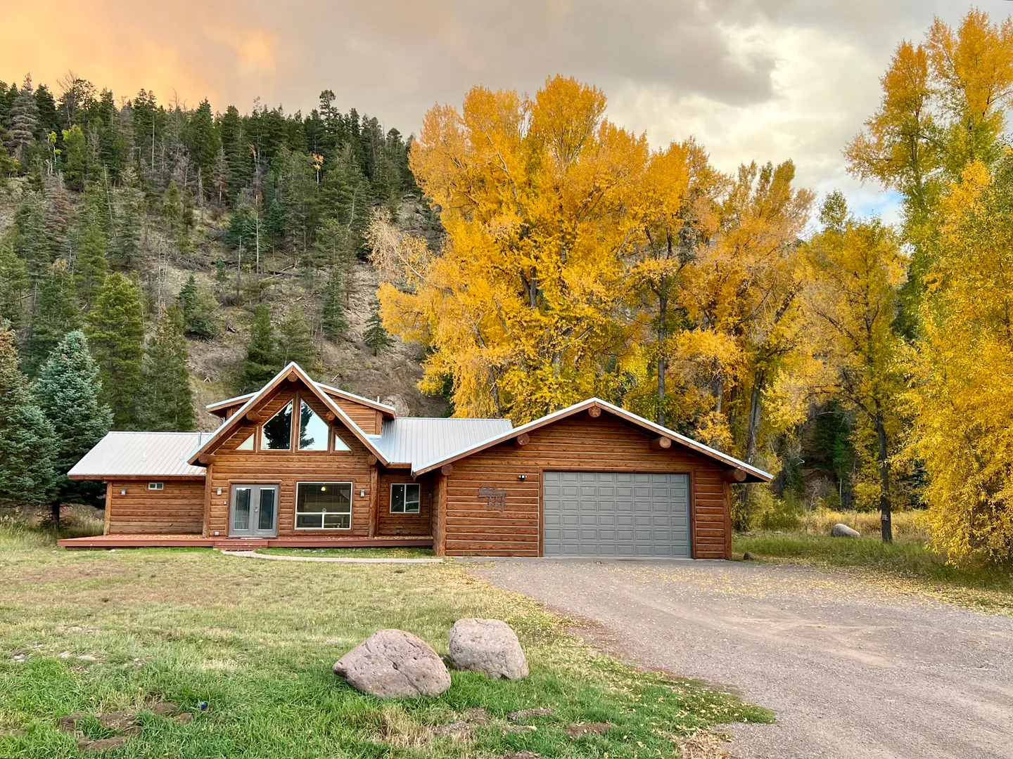 Exterior of a log-sided cabin with an attached garage, front deck, and large triangular gable window against a backdrop of autumn trees and a forested hillside.