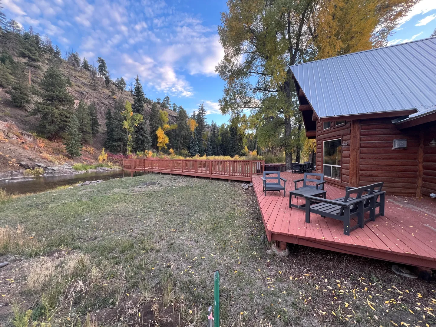 Outdoor wooden deck with seating area outside a log cabin next to a lawn and river flanked by pine-covered hills.