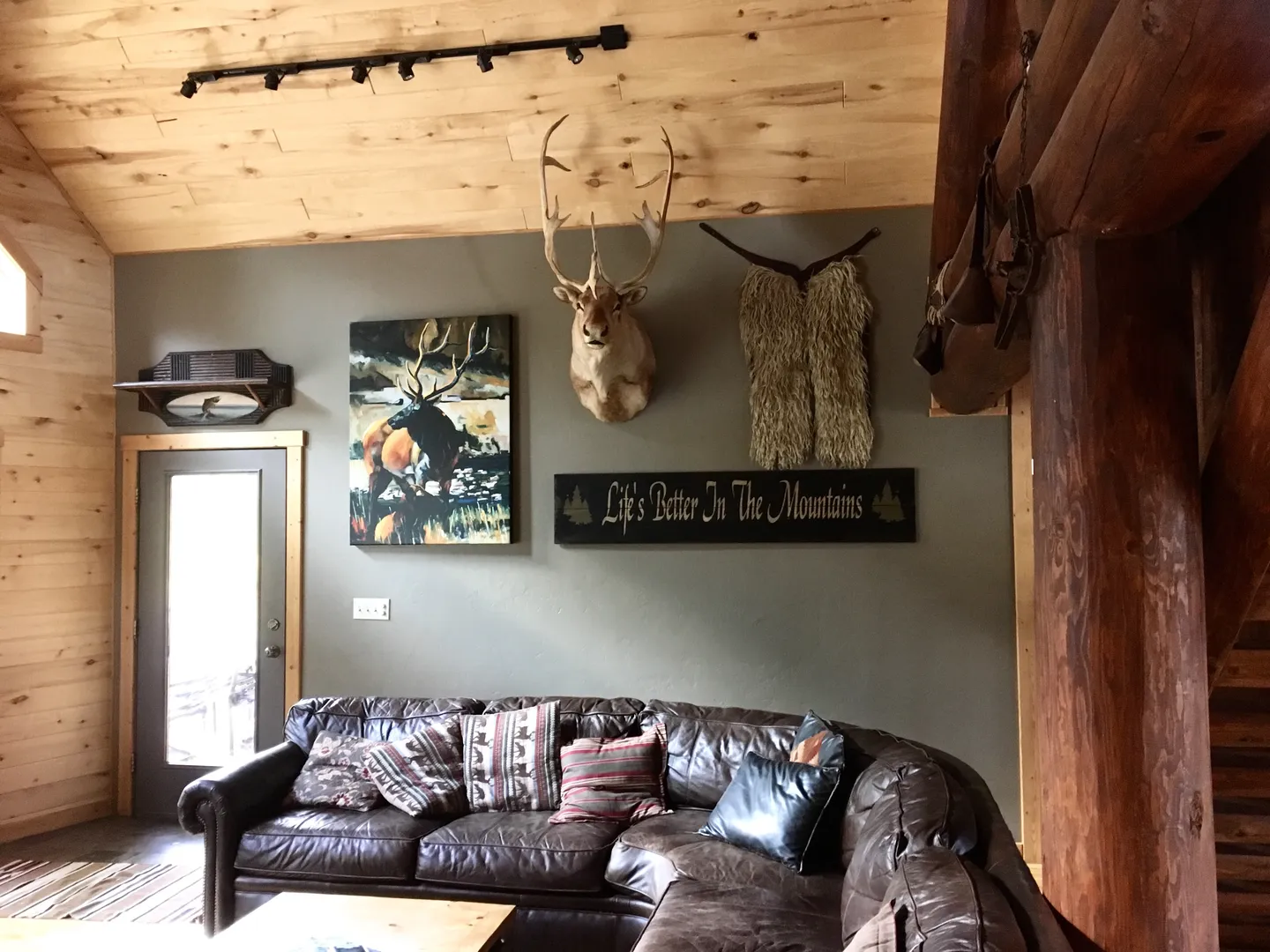 Living area with a brown leather sectional couch, wood-paneled ceiling, and rustic wildlife decor on the wall.