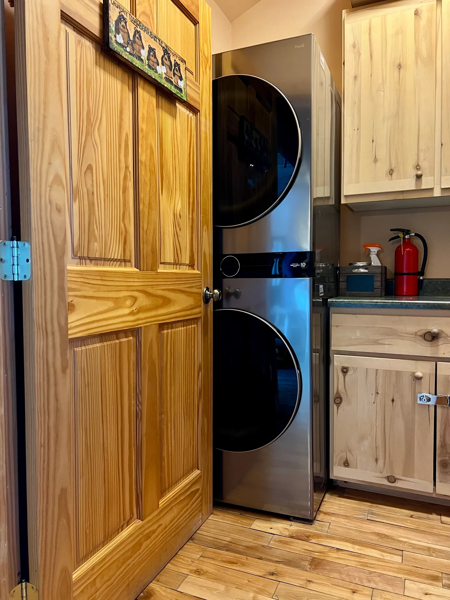 A stacked metallic front-load washer and dryer set tucked into a corner next to light wood cabinetry and a wood door.