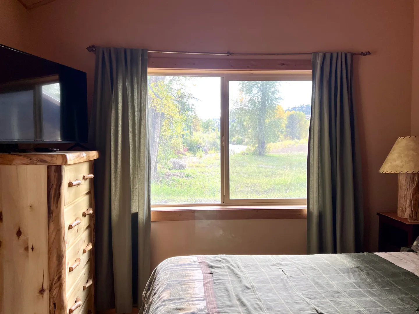 Bedroom window framed by curtains looking out onto an open green yard with trees.