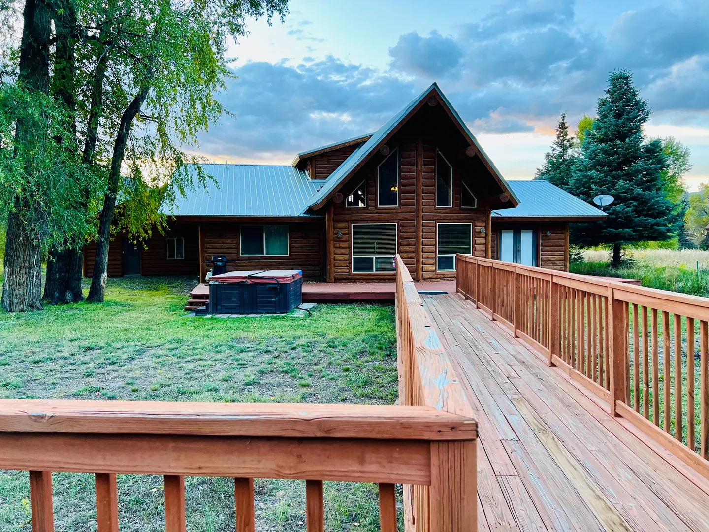 Exterior view of a log cabin with a metal roof, outdoor hot tub, and a wooden bridge walkway leading across the yard.
