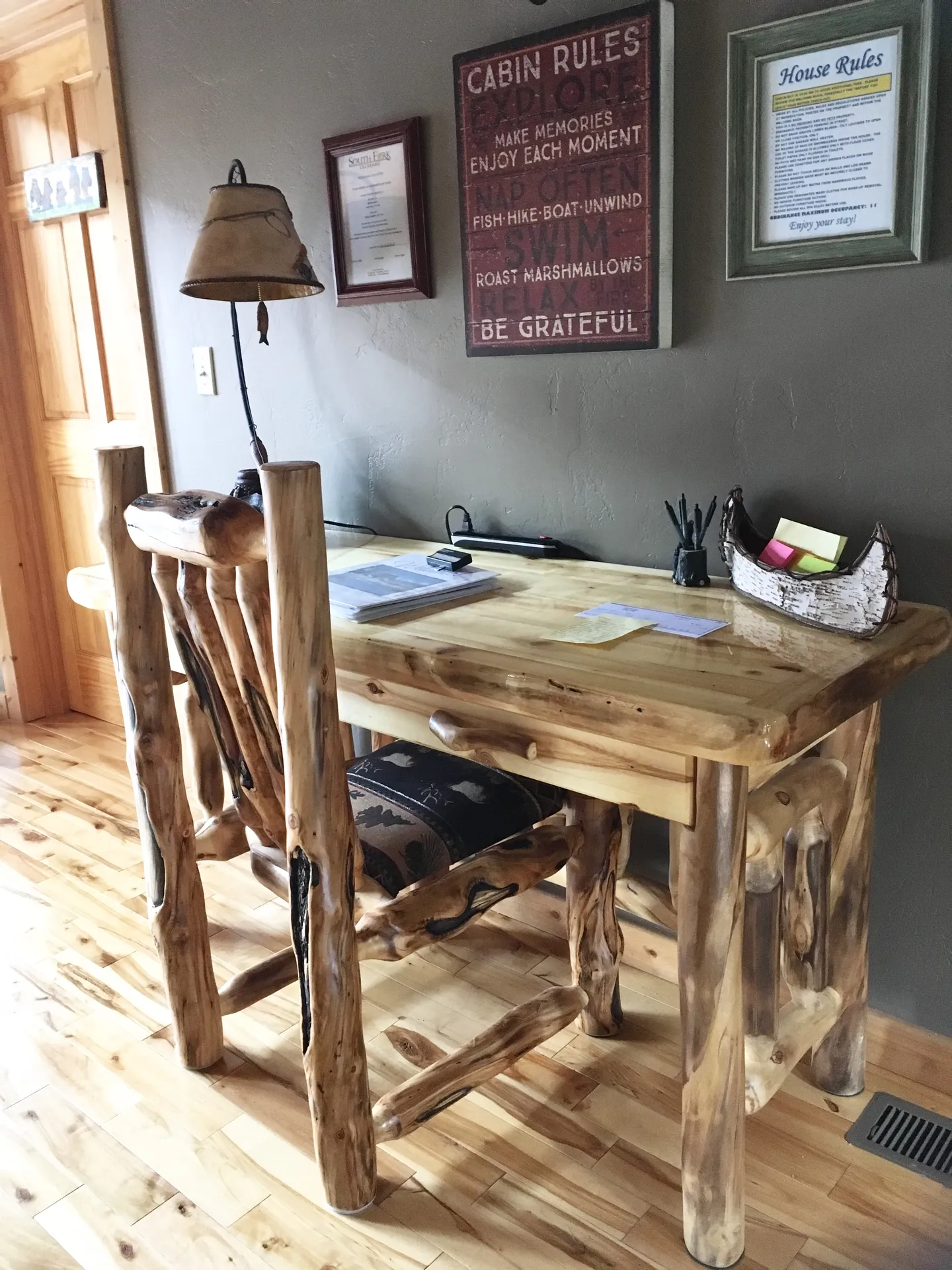 A rustic log-style desk and matching chair set against a gray wall with framed signs and light hardwood floors.