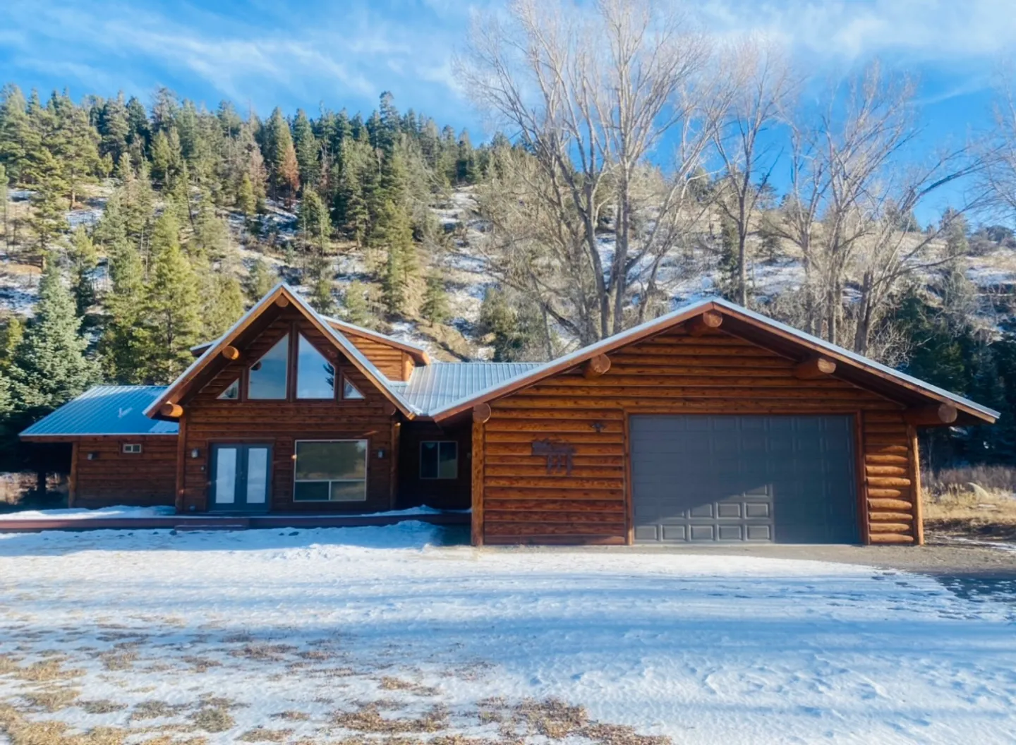 Exterior of a log home with an attached two-car garage set against a snow-dusted, forested hillside under a blue sky.