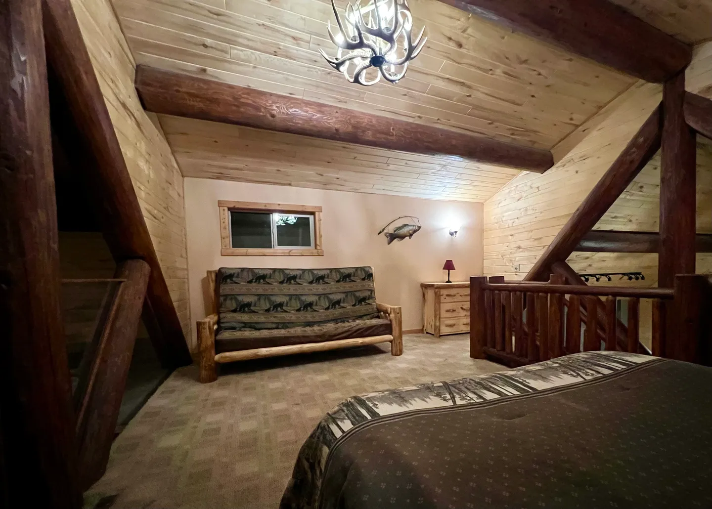 A rustic loft bedroom featuring wood-paneled angled ceilings, log beams, an antler chandelier, a futon couch, and part of a bed.