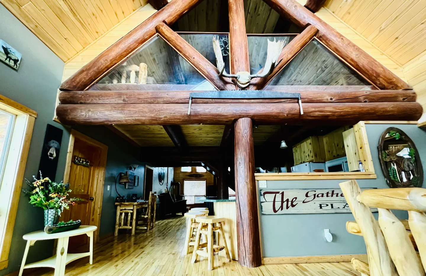 Open interior of a log cabin featuring heavy log beams, wood floors, an upper loft with a moose skull mount, and a view into the kitchen and dining area.