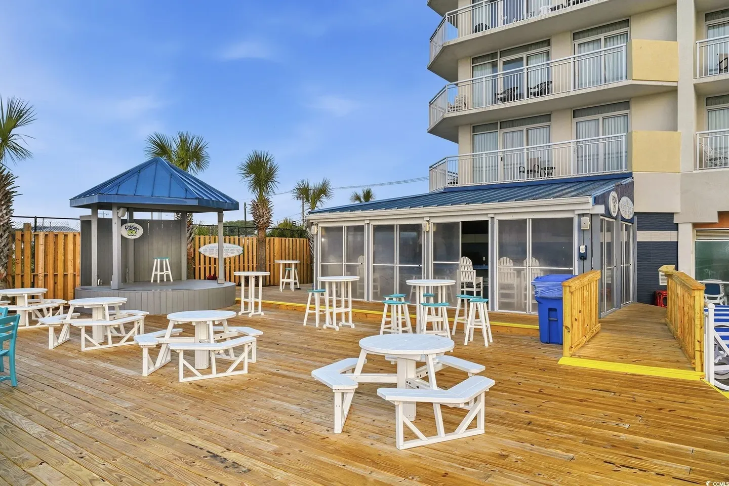 Outdoor wooden deck featuring picnic tables, tall bar tables with stools, a small covered stage, and a screened-in structure next to a multi-story building.