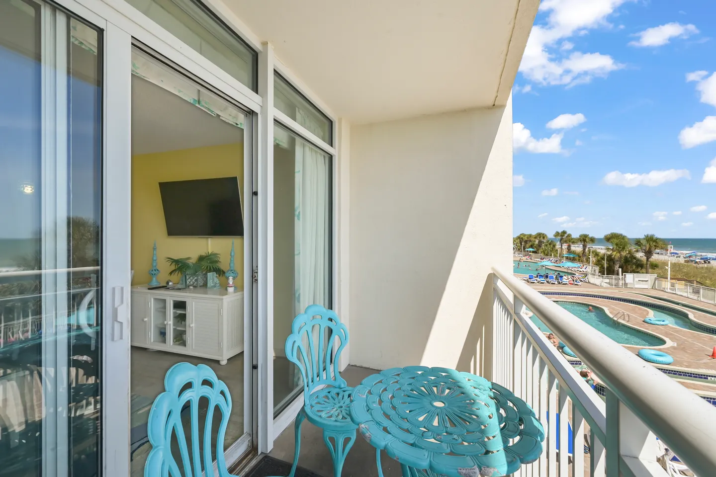Balcony with turquoise metal seating overlooking swimming pools and the ocean through sliding glass doors to the living area.