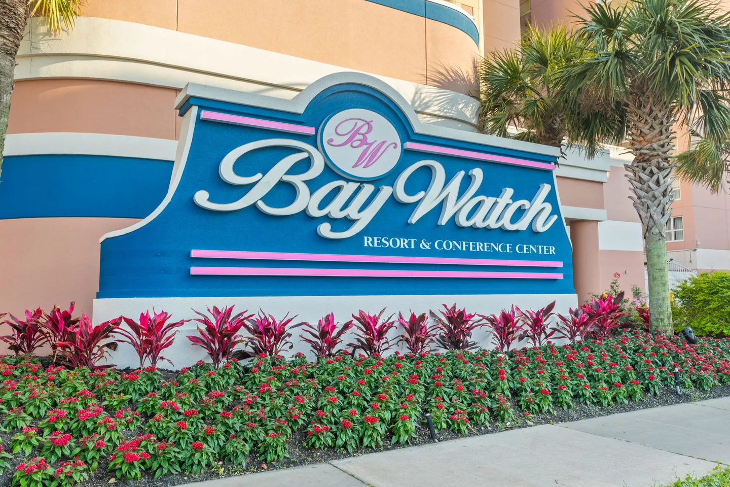 Exterior resort entrance sign reading Bay Watch Resort and Conference Center bordered by tropical plants and palm trees.