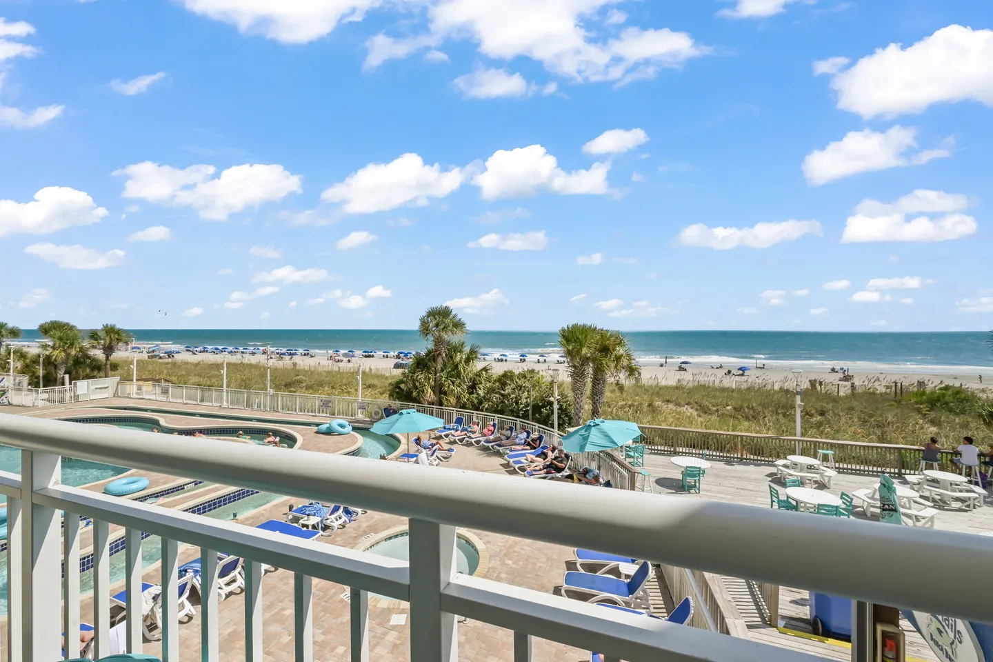 Balcony view overlooking a beachfront resort pool area, lazy river, palm trees, and the ocean under a blue sky.