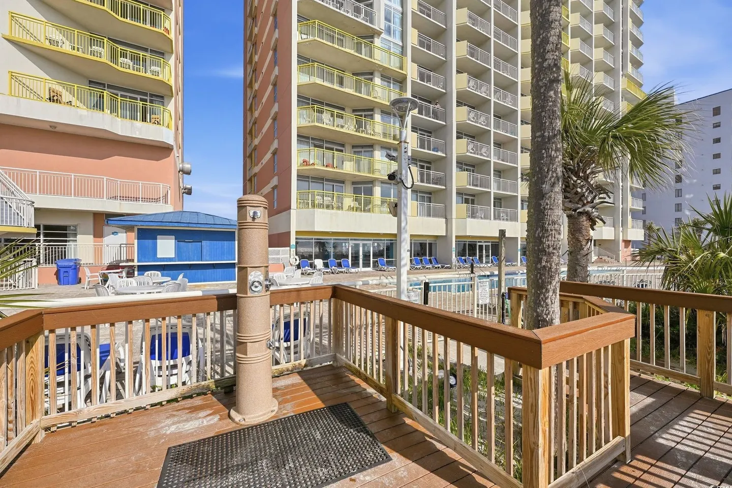 Outdoor shower station on a wooden boardwalk leading toward a multistory resort building and pool area.