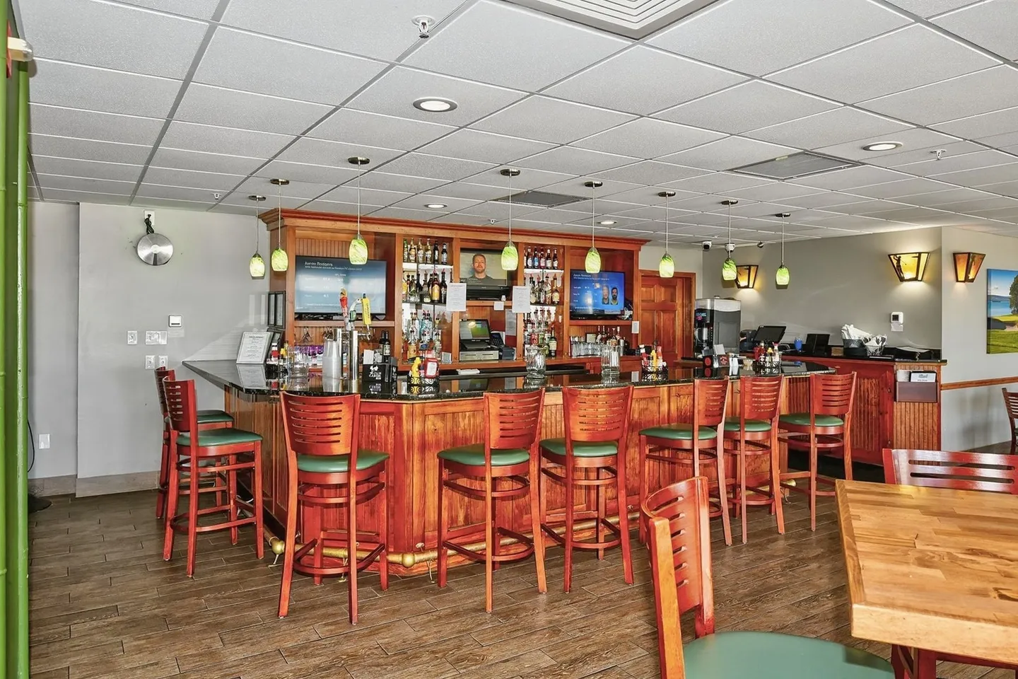 Commercial-style bar area with wooden paneling, a dark counter, green-cushioned barstools, and mounted TVs behind the counter.