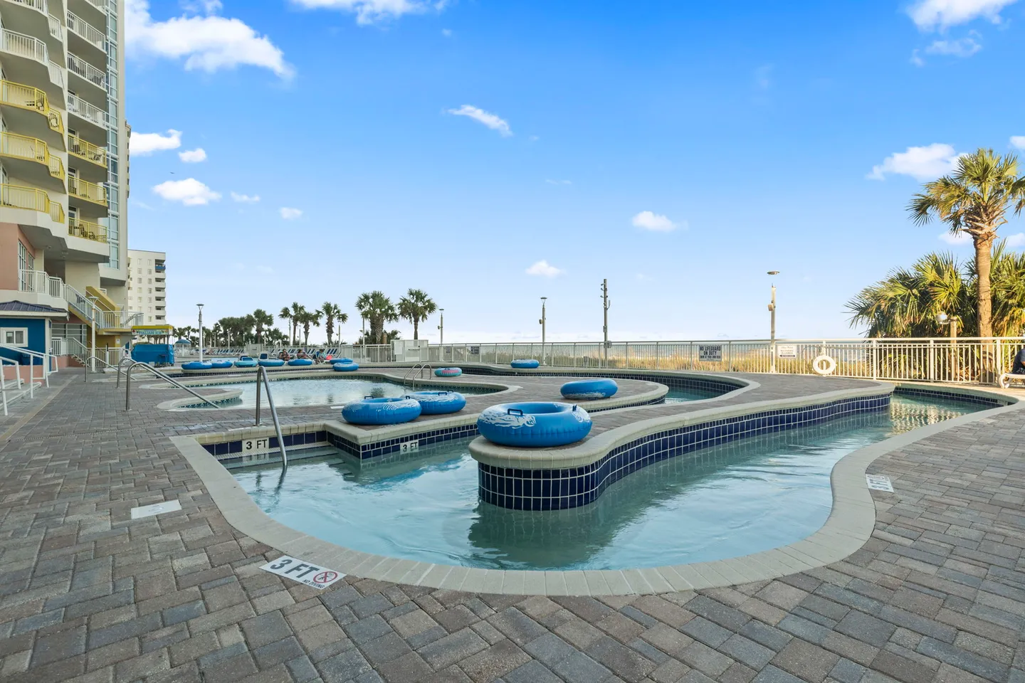 Outdoor lazy river pool with blue inner tubes surrounded by a paver deck, palm trees, and an oceanfront railing under a blue sky.