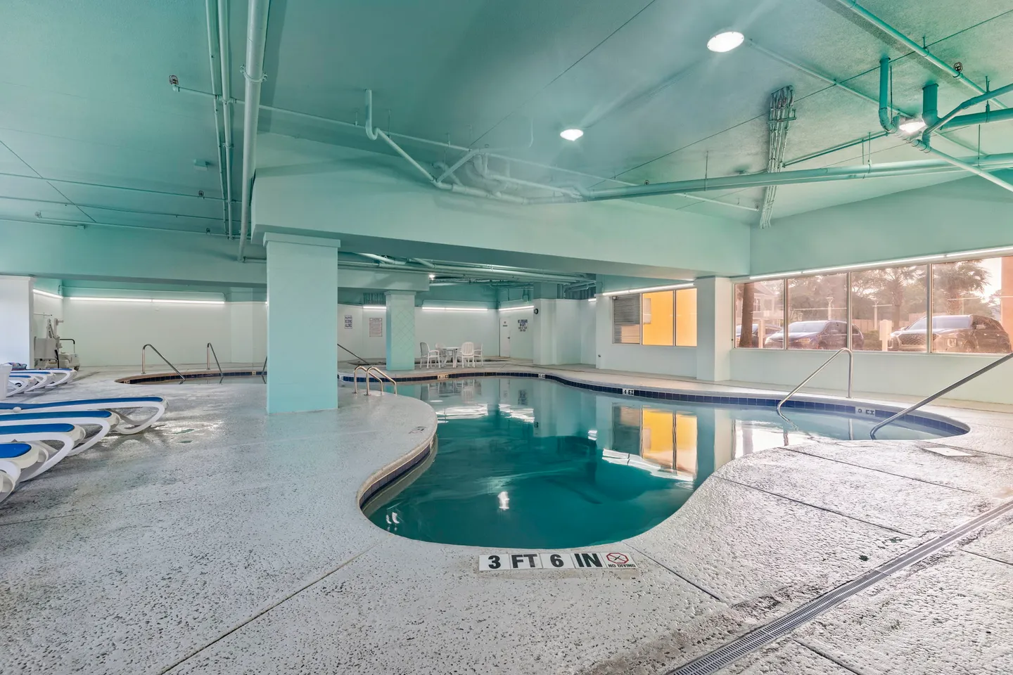 Indoor swimming pool with light aqua ceiling and columns, surrounded by lounge chairs and windows.
