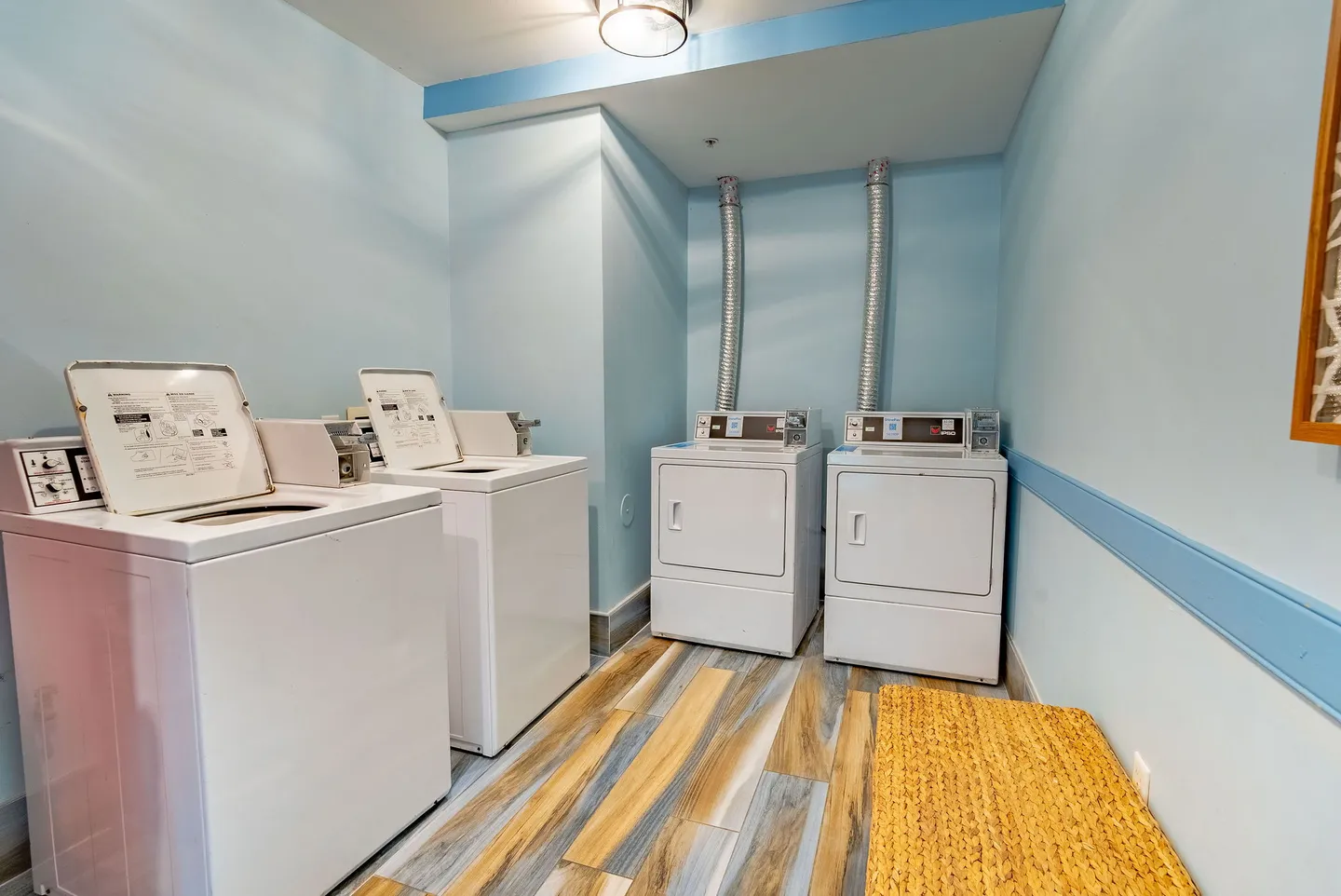A laundry room with light blue walls, wood-look plank flooring, two top-loading washing machines, and two front-loading dryers.