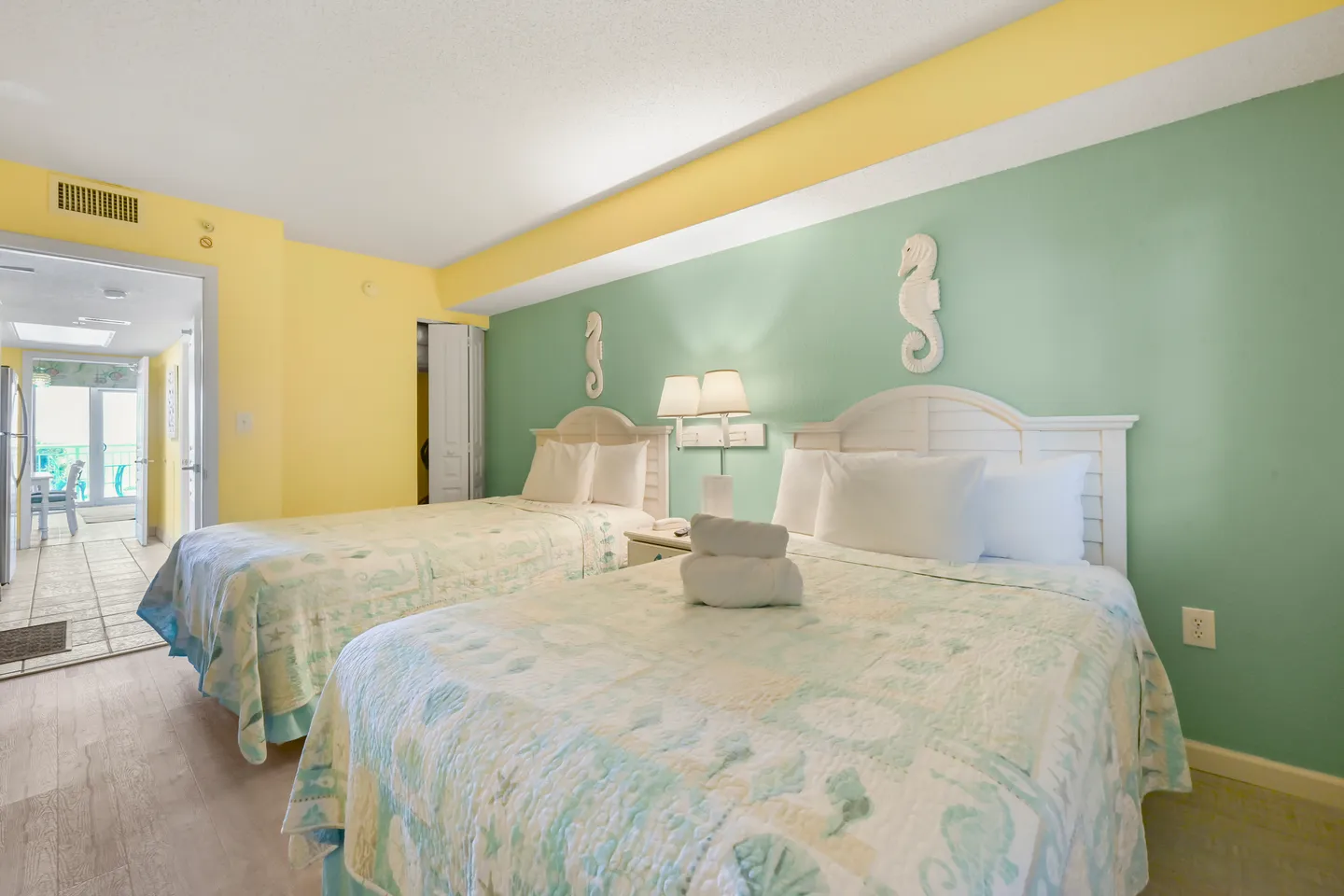 Bedroom with two beds, mint green and yellow walls, white headboards, and a doorway showing a balcony in the distance.