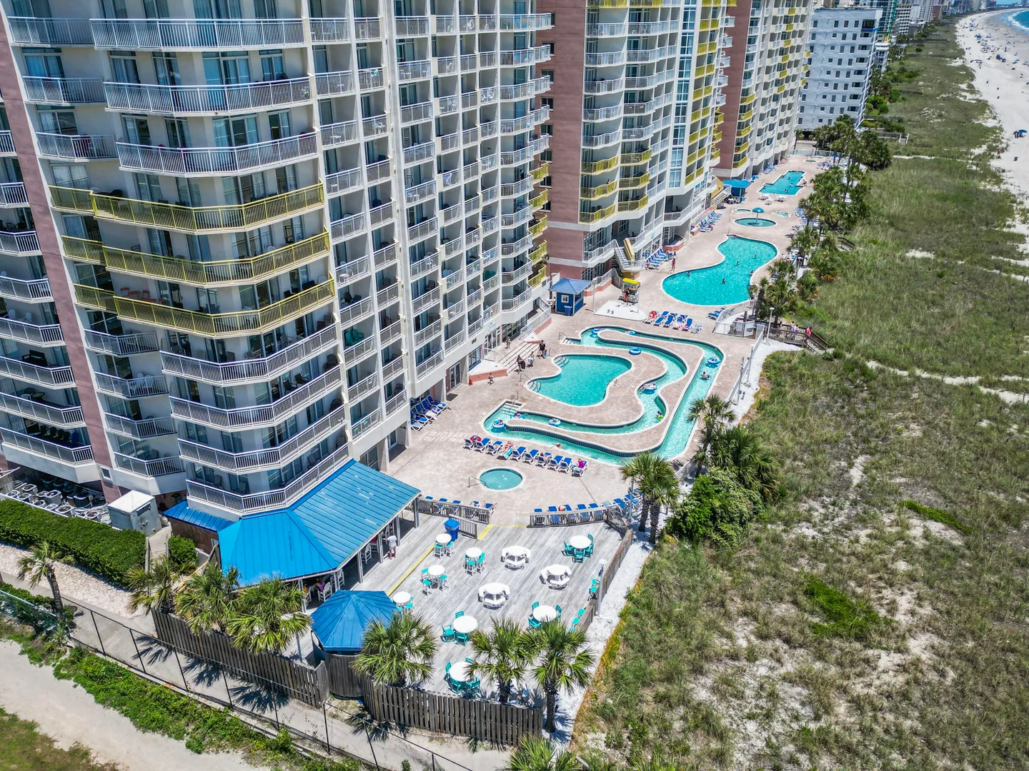 Aerial view of a beachfront high-rise resort featuring a lazy river, swimming pools, hot tub, and patio seating next to sand dunes.