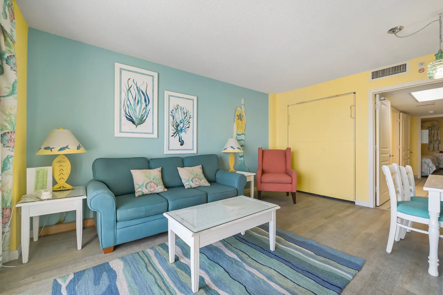 Living area with a turquoise sofa, white coffee table, striped blue rug, and coastal artwork on light blue and yellow walls.