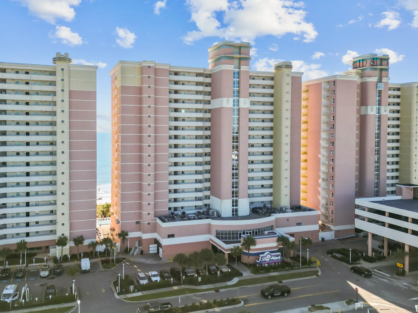 Aerial view of a tall, multi-story pink beachfront resort building with palm trees and a parking lot.