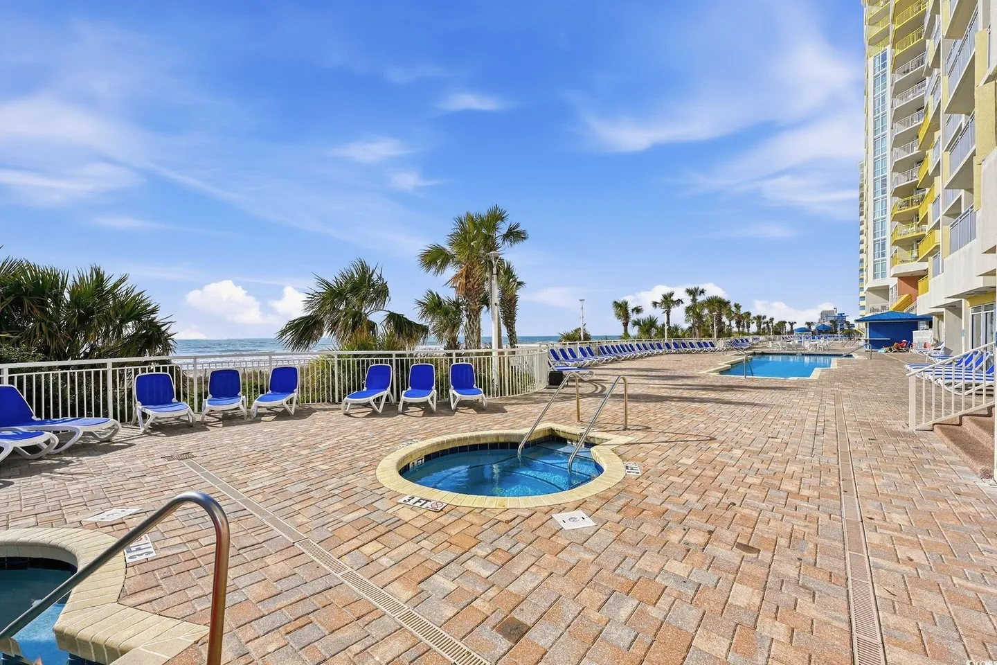 Outdoor paved pool deck with round hot tubs, lounge chairs, palm trees, and ocean views next to a multistory building.