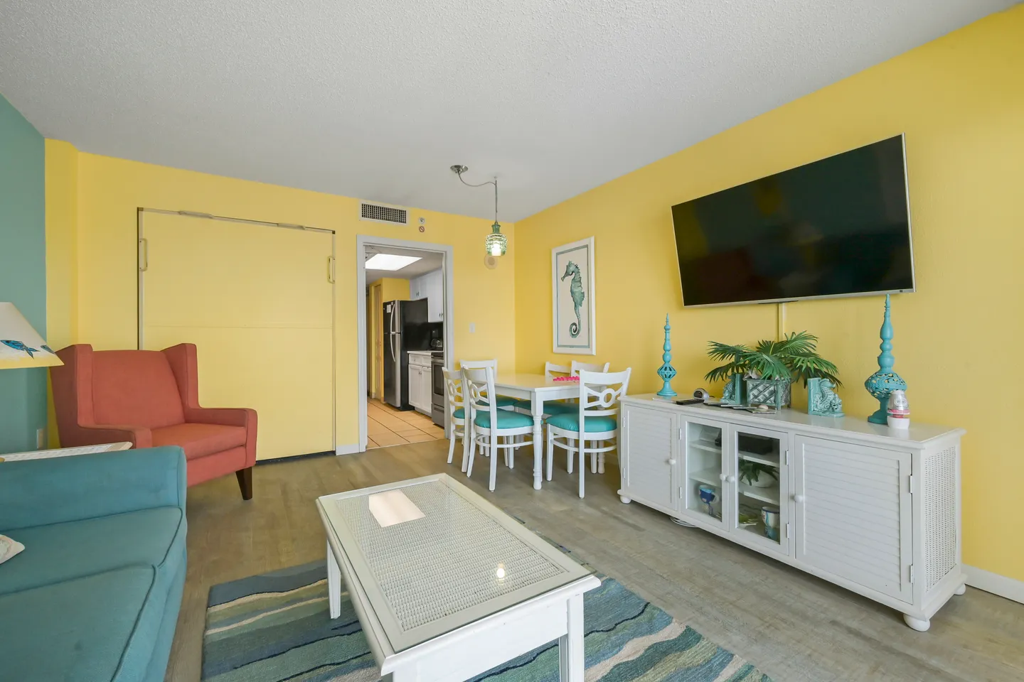 Living and dining area with yellow walls, a wall-mounted TV, dining table with chairs, coffee table, and an accent armchair.