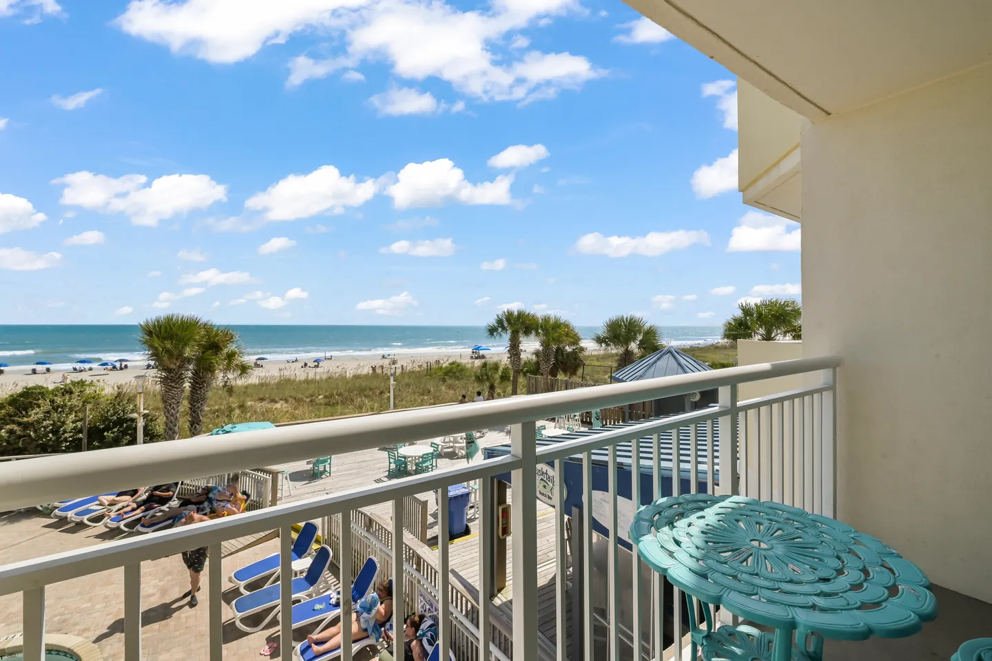 Elevated view from a balcony with white railings overlooking a pool deck, beach, and ocean under a blue sky with clouds.