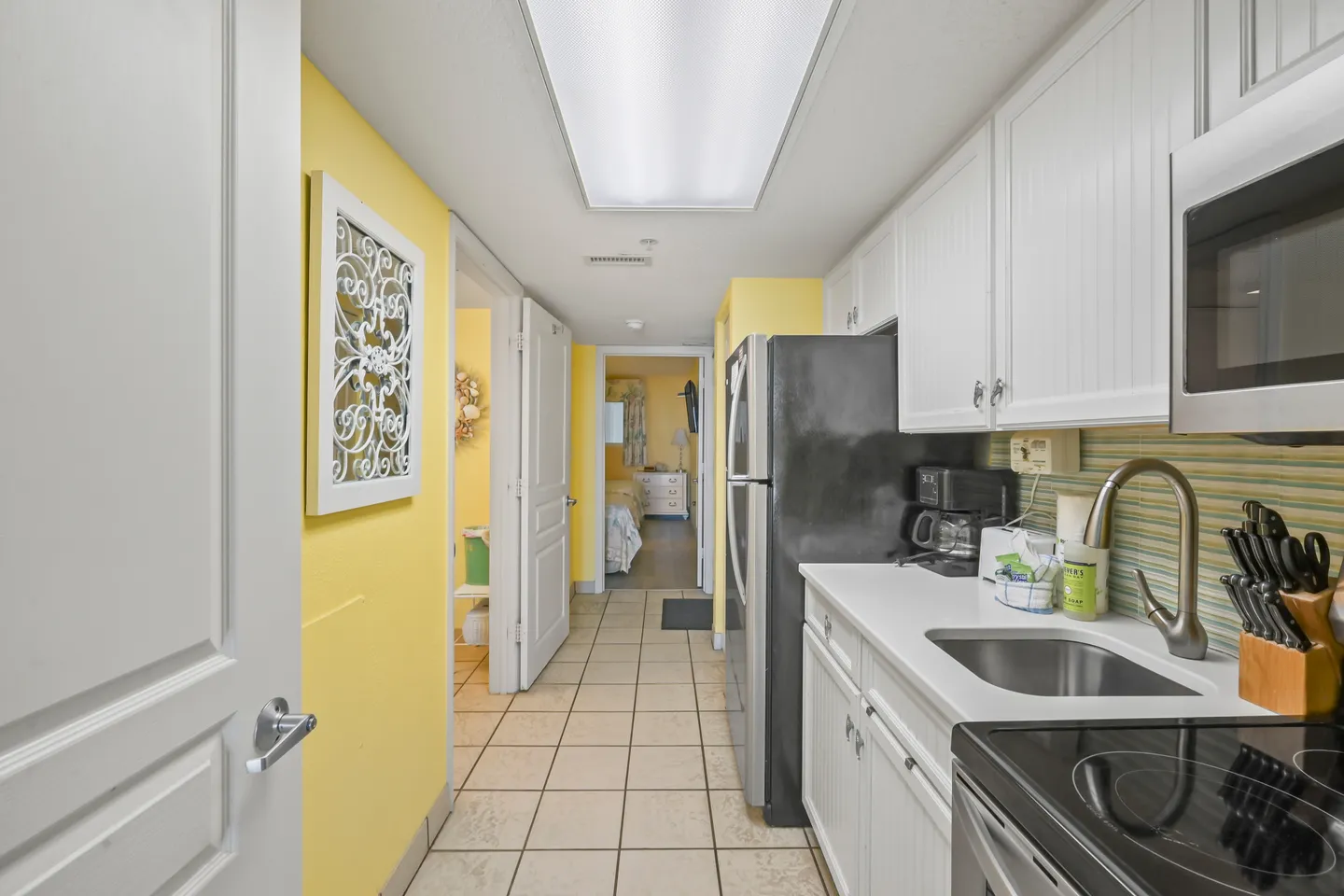 Galley kitchen with white beadboard cabinets, light countertops, appliances, and a tiled hallway leading to bedrooms and a bathroom.