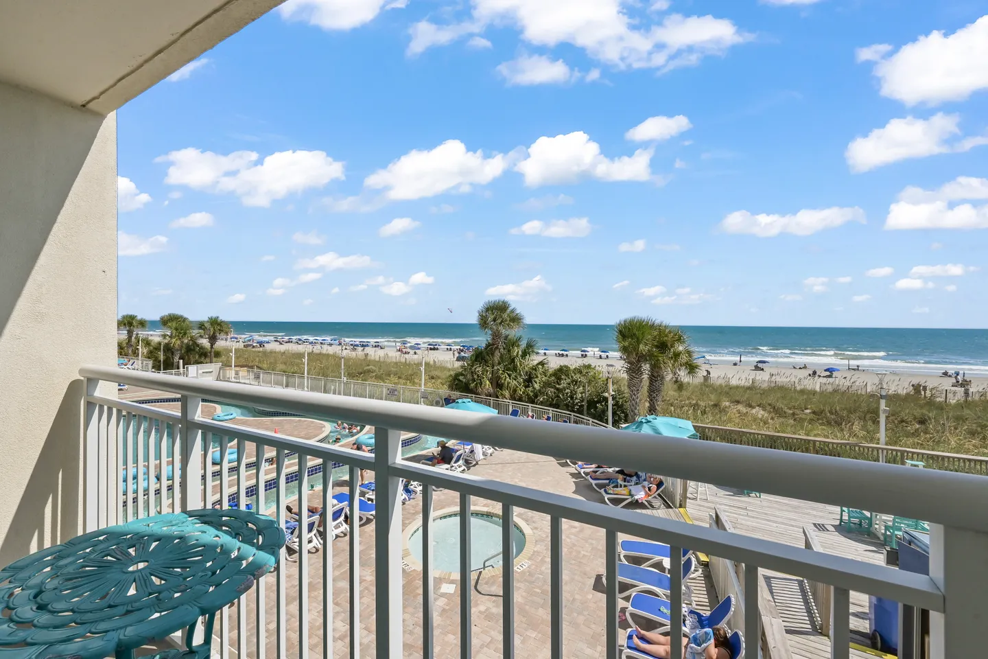 Balcony view overlooking a resort pool, hot tub, palm trees, sandy beach, and ocean under a blue sky.