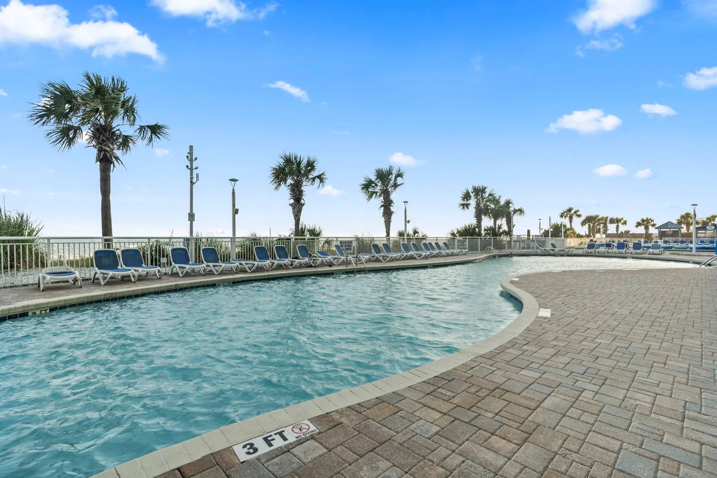 Outdoor swimming pool surrounded by paved sun decks, lounge chairs, and palm trees under a bright blue sky.
