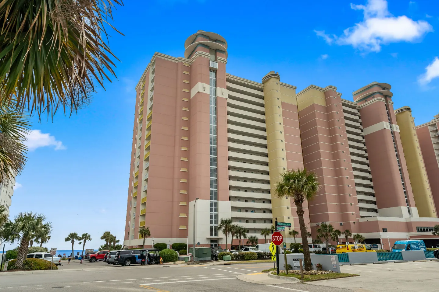 Exterior view of a multi-story pink and beige high-rise condominium complex near the street and palm trees.