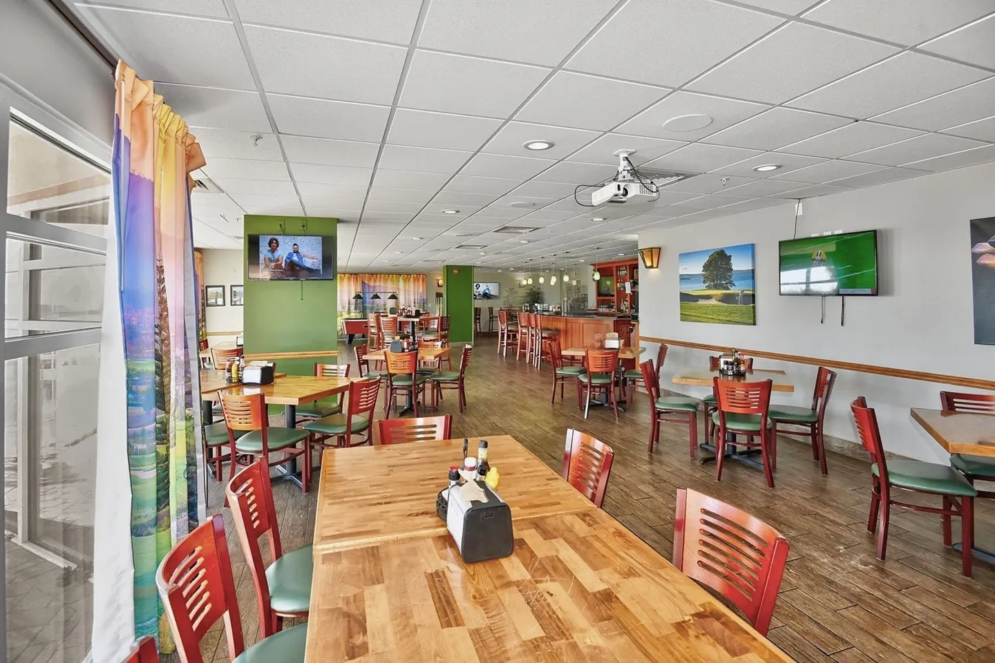 Spacious dining area featuring multiple wood tables with red and green chairs, wood-look flooring, wall-mounted TVs, and a ceiling-mounted projector.