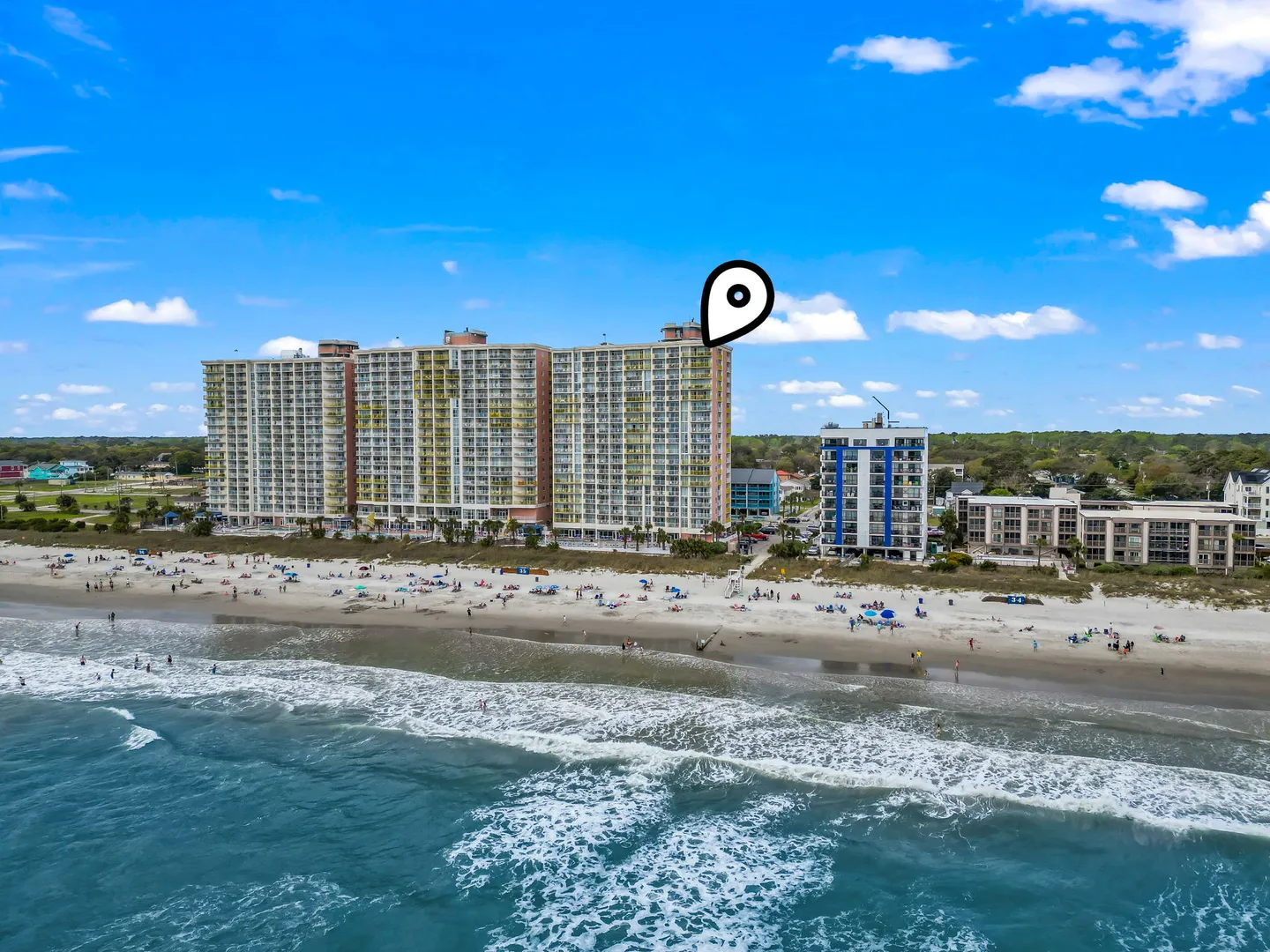 Aerial view of an oceanfront high-rise building on a sandy beach next to the blue ocean under a partly cloudy sky.
