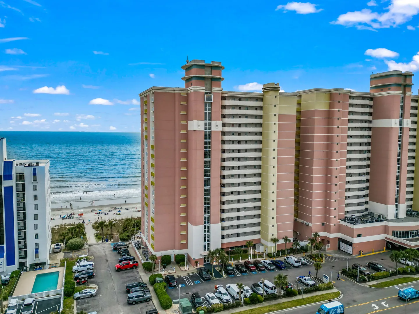 Aerial view of a tall, pink beachfront high-rise resort building with parking lots and the ocean behind it under a blue sky.