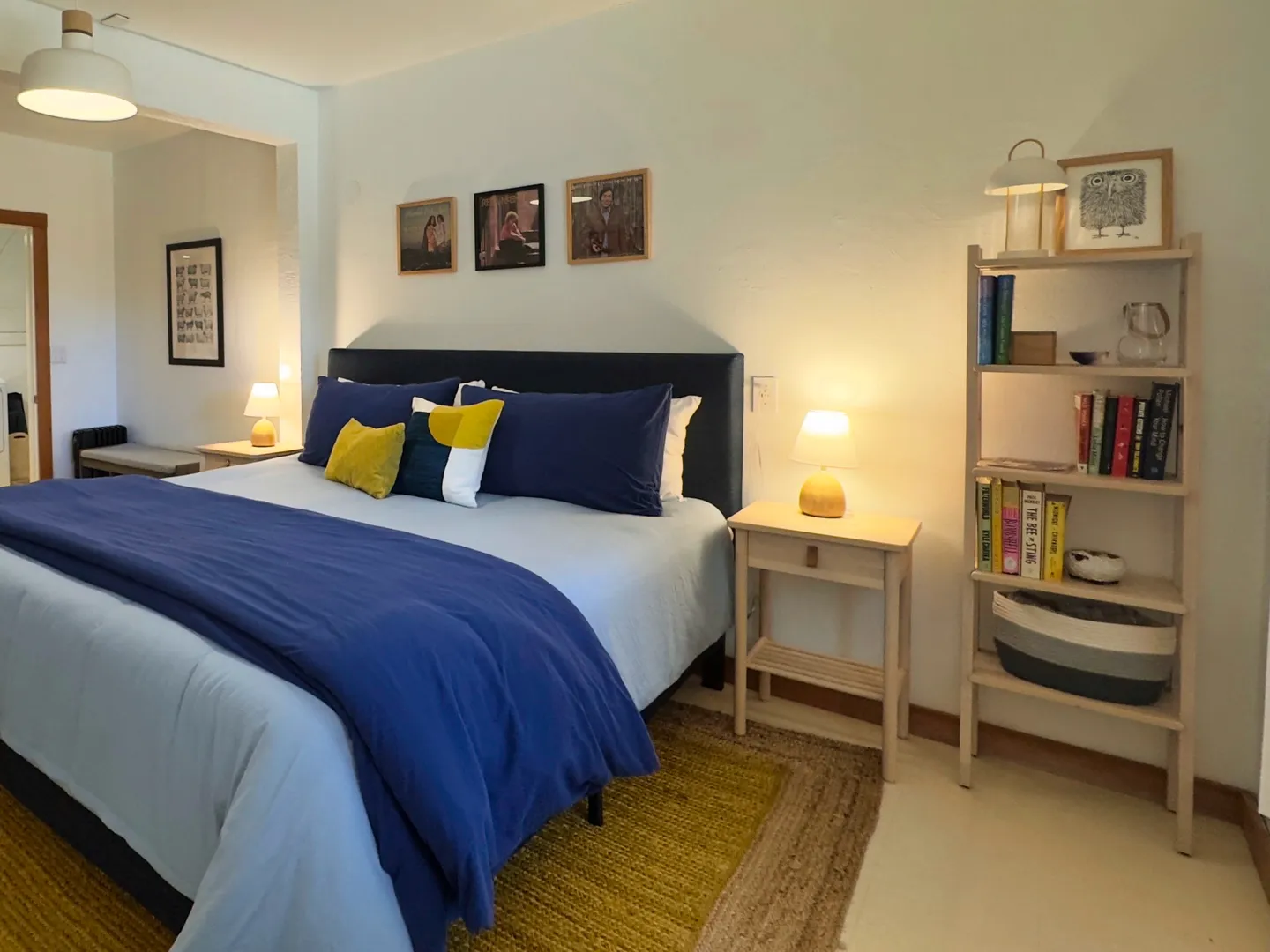 Bedroom with a large bed dressed in blue and white linens, a nightstand with a lamp, and a tall light-wood bookshelf.