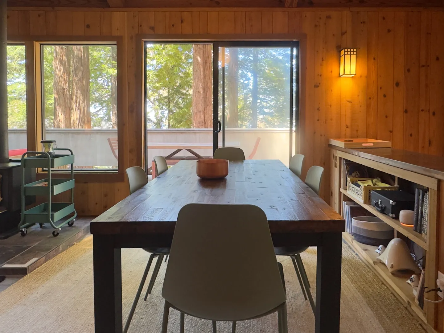 Dining room with a long wood table surrounded by chairs, wood-paneled walls, and sliding glass doors looking out to a deck and trees.