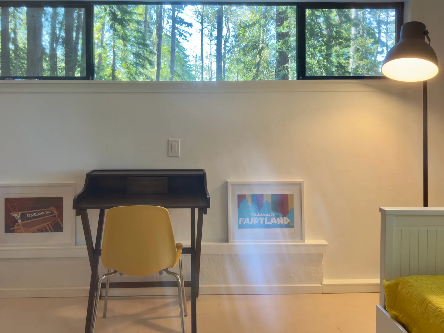 A small writing desk with a yellow chair beneath a long horizontal window looking out to trees.