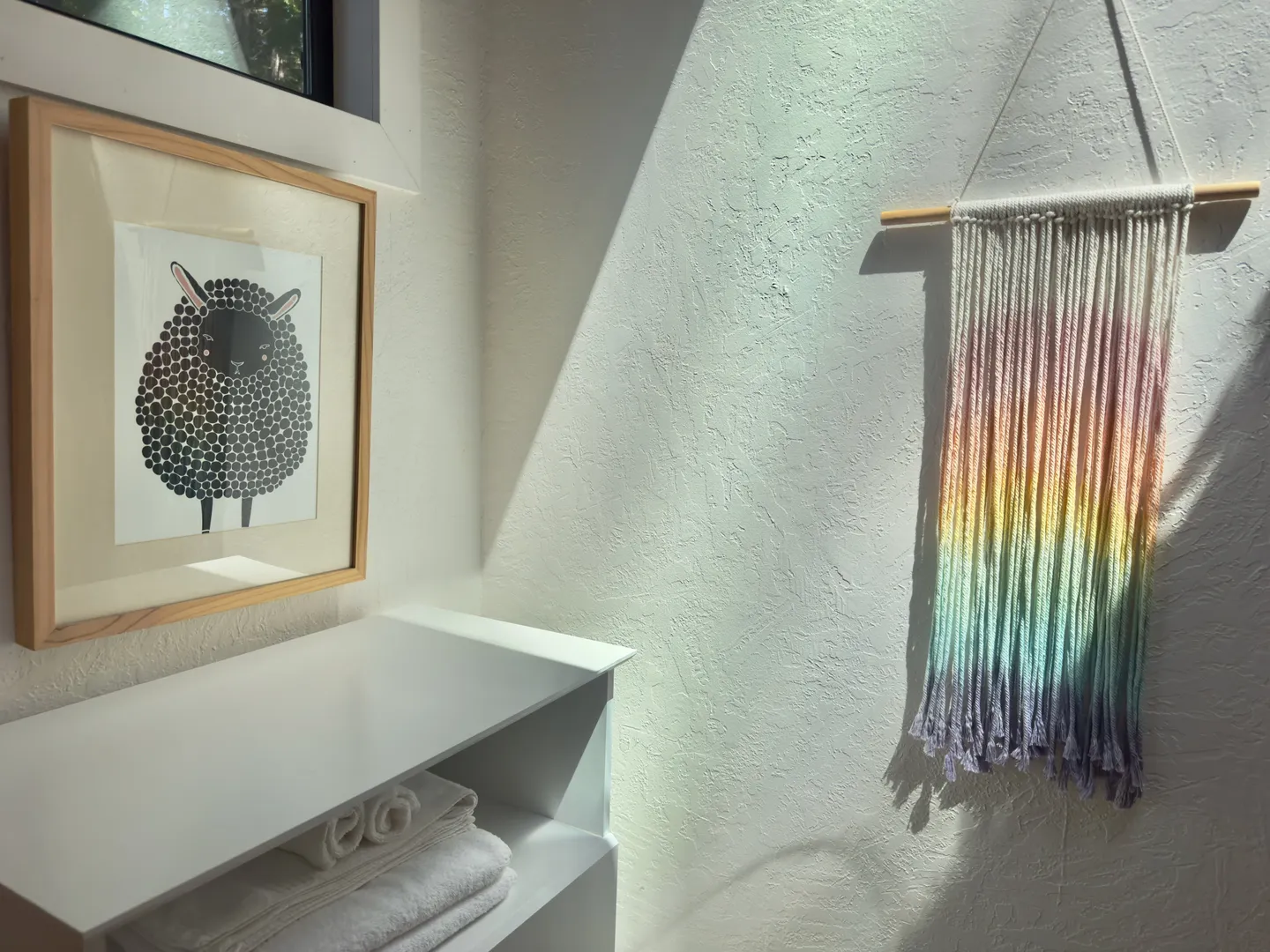 Corner of a room with textured white walls, a framed sheep print, a rainbow macrame wall hanging, and stacked white towels on a shelf.