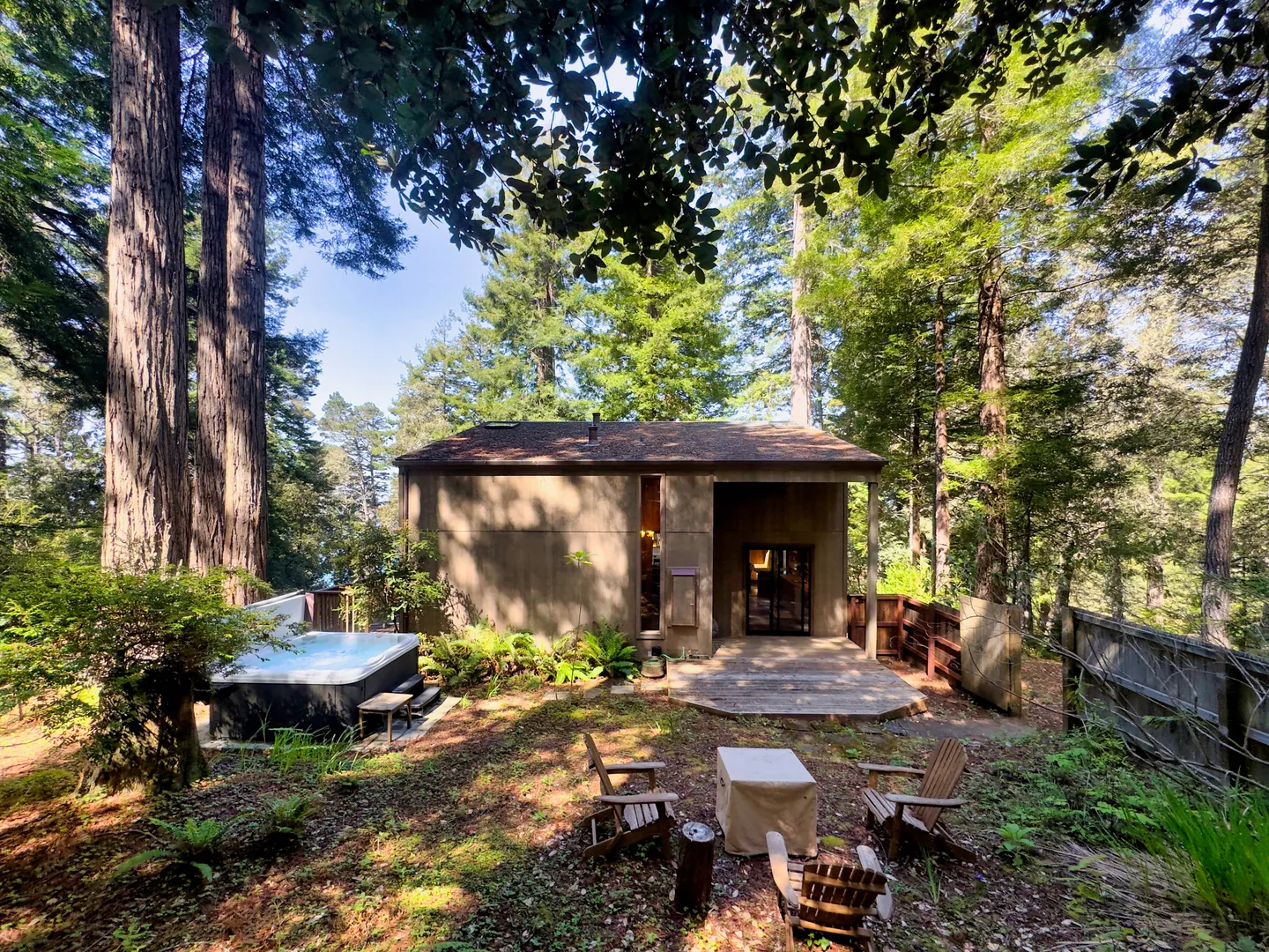 Exterior backyard view of a wooden cabin in a redwood forest with a hot tub, deck, and outdoor fire pit area.