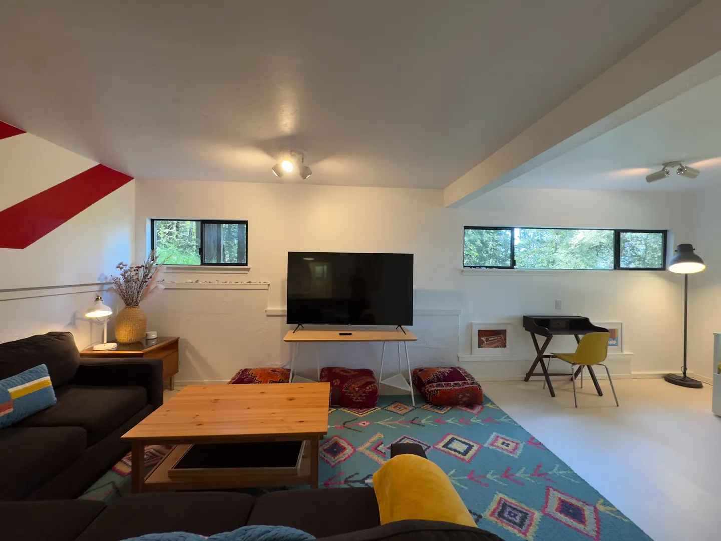 Living room featuring a dark sofa, light wood coffee table, colorful geometric rug, television on a stand, floor cushions, and a small desk in the corner.